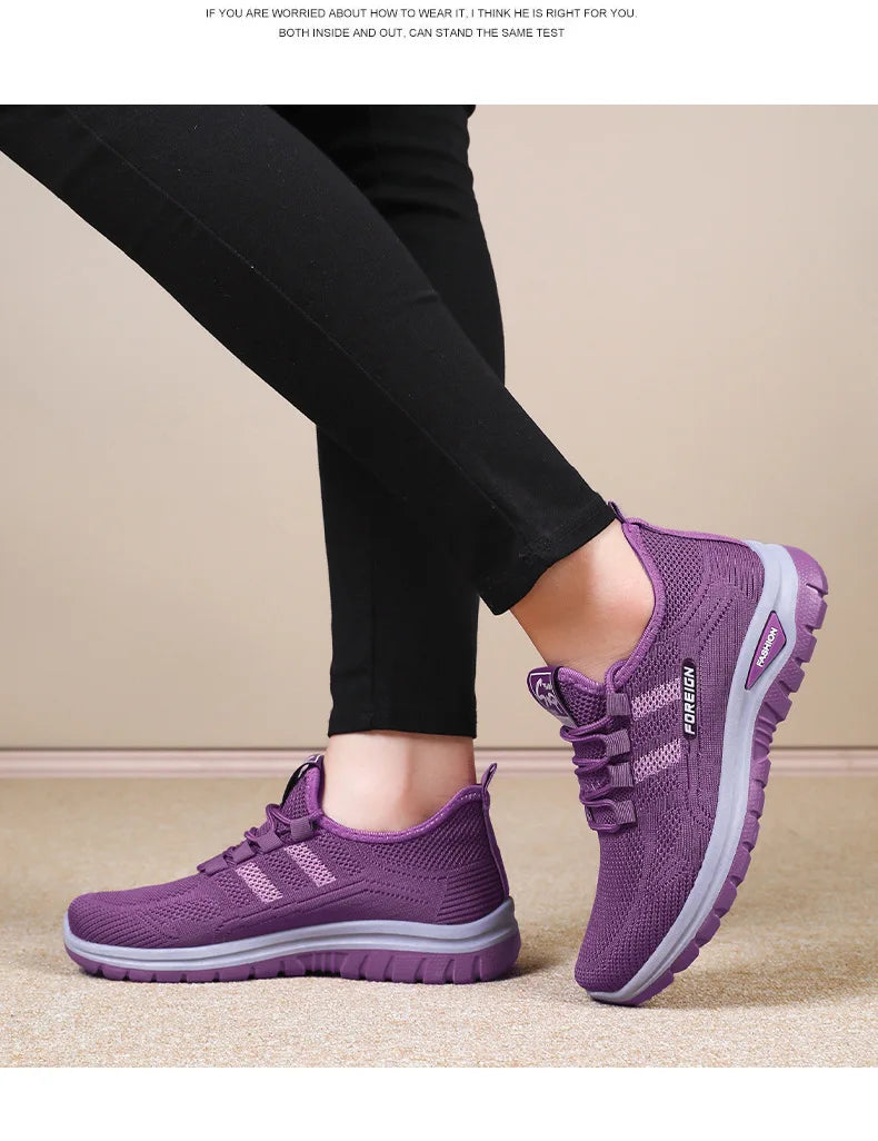 A person wearing black pants models purple mesh running shoes with grey soles indoors.
