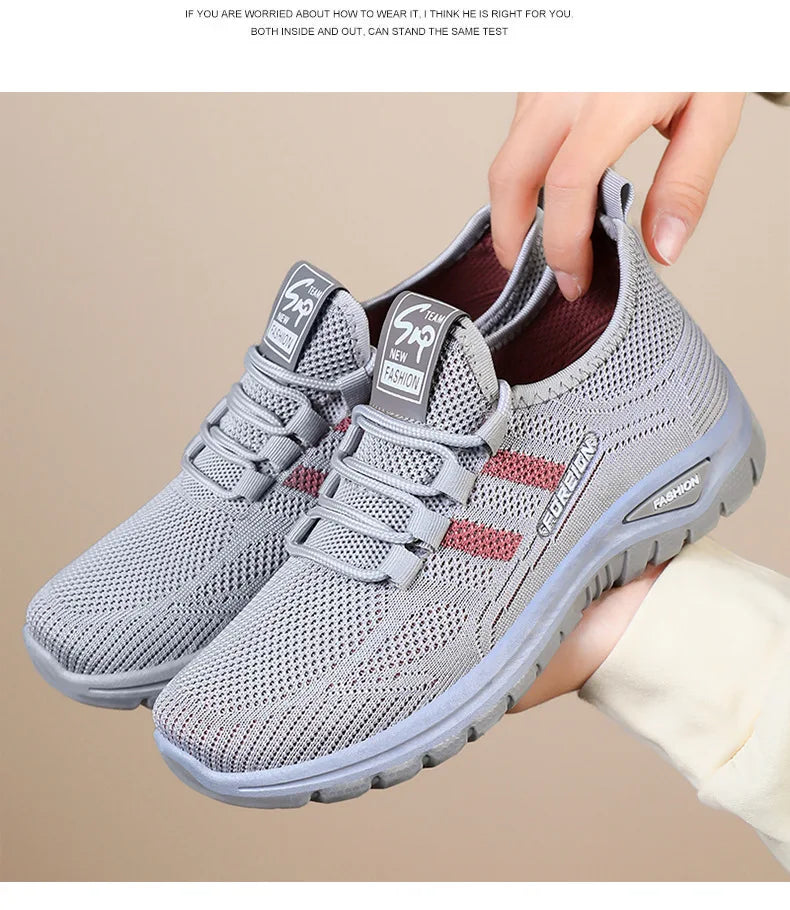 Hands display a pair of lightweight gray mesh running shoes with red accents, showcasing breathable fashion footwear.
